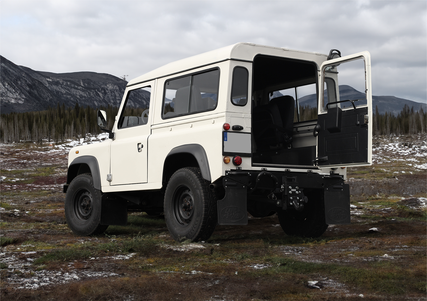 Heckansicht eines weißen restaurierteren Land Rover Defender 90 in rauer alpinen Landschaft. Berge und Bäume im Hintergrund