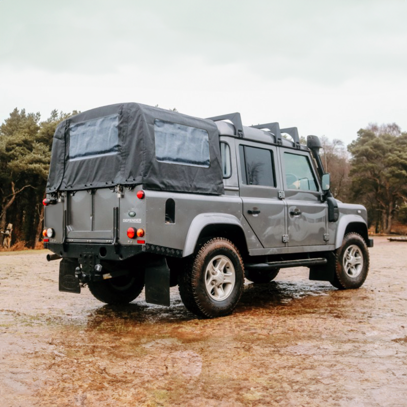 Land Rover Defender 110 Station Wagon Crewcab in grau metallic mit PVC Verdeck in schwarz auf einer verregneten Wiese mit Wald im Hintergrund