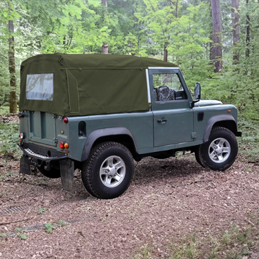 Land Rover Defender 90 mit Soft Top Verdeck in oliv grün aus Canvas im Wald. Im Hintergrund sind Bäume zu sehen. Das Verdeck hat ein Heckfenster.
