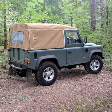 Land Rover Defender 90 mit Soft Top Verdeck in sand beige aus Canvas im Wald. Im Hintergrund sind Bäume zu sehen. Das Verdeck hat ein Heckfenster.