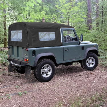 Land Rover Defender 90 mit Soft Top Verdeck in schwarz aus Canvas im Wald. Im Hintergrund sind Bäume zu sehen. Das Verdeck hat seitliche Fenster und ein Heckfenster.