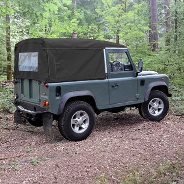 Land Rover Defender 90 mit Soft Top Verdeck in schwarz aus Canvas im Wald. Im Hintergrund sind Bäume zu sehen. Das Verdeck hat ein Heckfenster.